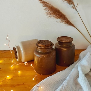 Könnte beinhalten: Drei Keramik-Gläser mit Korkdeckeln, zwei braun und eines weiß, auf einer Holzoberfläche mit einem weißen Tuch und Lichterketten.