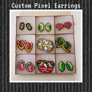 Puede incluir: Un conjunto de ocho pares de pendientes de arte de píxeles en varios diseños, que incluyen corazones, pociones y personajes de videojuegos. Los pendientes se muestran en pequeñas cajas con fondos blancos.