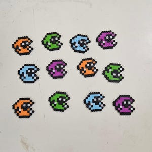Puede incluir: Una colección de figuras de pixel art coloridas, cada una parecida a un fantasma o personaje estilizado. Las figuras son de varios colores, incluyendo naranja, verde, azul y morado, con contornos negros. Están dispuestas sobre una superficie clara.