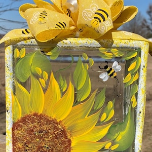 May include: A glass block with a painted sunflower and a bee, topped with a yellow bow with a bee pattern.