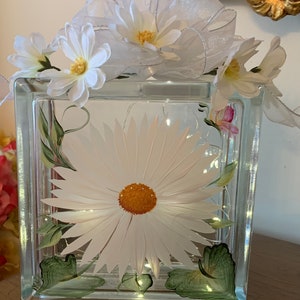 May include: A clear glass block with a painted white daisy and green leaves. The block is decorated with a white sheer ribbon and white daisies.