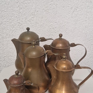 Peut inclure: Un ensemble de quatre cafetières métalliques de tailles variées, chacune dotée d'un corps arrondi, d'une anse courbée, d'un bec verseur et d'un couvercle avec un petit bouton. Les pots présentent une finition en cuivre et laiton patiné.
