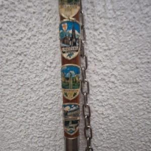 Könnte beinhalten: Ein Vintage-Souvenirstock mit mehreren dekorativen Schilden mit farbenfrohen Stadtlandschaften und Wahrzeichen. Eine silberne Kette hängt an dem Stock. Der Stock ist braun mit silbernen Akzenten.