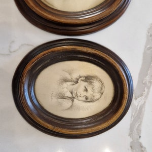Könnte beinhalten: Zwei ovale gerahmte Bleistiftzeichnungen von Kindern. Die Rahmen sind aus dunkelbraunem Holz mit einem helleren braunen Innenrand.