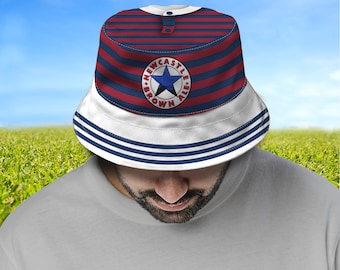 Puede incluir: Un sombrero de pescador con una corona a rayas rojas y azul marino y un ala blanca. El sombrero presenta un emblema circular con una estrella azul y las palabras "Newcastle Brown Ale". El sombrero lo lleva una persona.