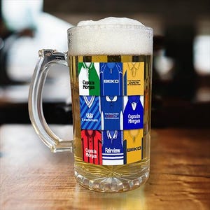 May include: A clear glass beer stein with a handle, filled with a light yellow liquid. The stein has a collage of colourful football shirts with team names and logos, including "Captain Morgan", "BEKO", "Lewisham", "Fairview", and "LIVETV".