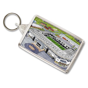 May include: A rectangular keyring with a detailed illustration of a stadium. The stadium has a green pitch, black and white seating, and a complex roof structure. The keyring has a silver ring.