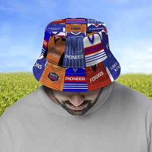 May include: A patchwork bucket hat with panels displaying sports jersey designs. The hat features the words "PIONEER", "FISONS", "MAGICAL VEGAS", and "GREENE KING". The hat is set against a blue sky and green grass background.