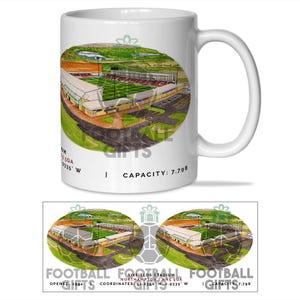 May include: White ceramic mug with a detailed illustration of a stadium. The image shows a green pitch, stands, and surrounding landscape. Text on the mug indicates the stadium's name, location, and capacity. A football-themed gift.