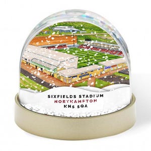 May include: A snow globe showing Sixfields Stadium, Northampton, England. The illustration depicts the stadium with green pitch, red seating, and surrounding buildings. The base of the globe displays the stadium's name and address. The globe is filled with white, glittery snow.