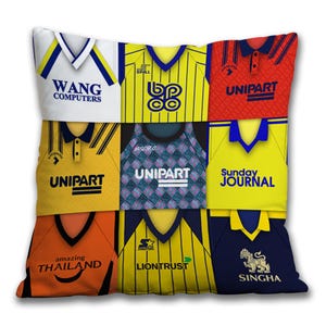 May include: Square cushion with a patchwork design of retro football shirts. The cushion showcases a variety of team jerseys in colours including white, yellow, red, orange, and navy, each with distinct logos and text such as "WANG COMPUTERS" and "SINGHA".