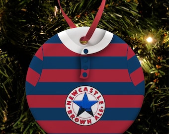 Puede incluir: Un adorno navideño rojo y azul a rayas con forma de camisa con cuello blanco y botón azul. El adorno tiene un círculo blanco con una estrella azul y el texto "NEWCASTLE BROWN ALE" en él.