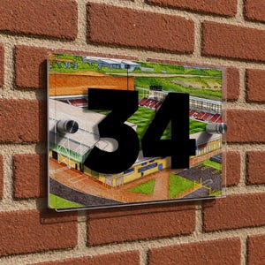 May include: A rectangular house number sign with a stadium illustration. The black number "34" is mounted on a clear acrylic panel with silver fixings. The sign is attached to a red brick wall. The stadium illustration is in watercolor.