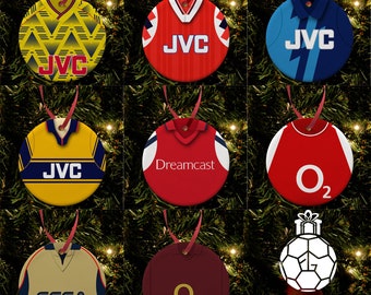 May include: Nine round Christmas ornaments featuring different Arsenal football jerseys from various seasons. The jerseys feature sponsors like JVC, Dreamcast, and O2. The ornaments are hanging on a Christmas tree. The bottom of the image reads "FOOTBALL GIFTS".