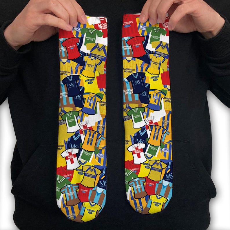 May include: A pair of colourful socks with a pattern of vintage football shirts. The socks are yellow, blue, red and white with the text "Mansfield Bitter" and "Evisons" repeated throughout the pattern.