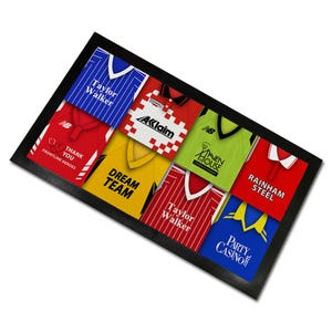 May include: A framed collage of nine different football shirts, each with a unique design and text. The shirts are in various colours, including red, blue, yellow, green, and white. The text on the shirts includes "Taylor Walker", "Dream Team", "Rainham Steel", "Party Casino", "Thank You Frontline Heroes", and "AKlaim".