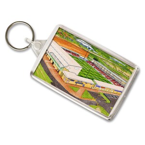 May include: A rectangular keyring with a watercolour illustration of a stadium, showing a green pitch, red seating, and tan buildings. The keyring has a silver ring attached.