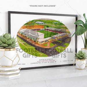 May include: A framed print depicting Sixfields Stadium, Northampton, with an illustrated view of the football ground. The artwork shows the stadium's design and surrounding areas. Text on the print reads "FOOTBALL GIFTS". The print is displayed on a white surface.