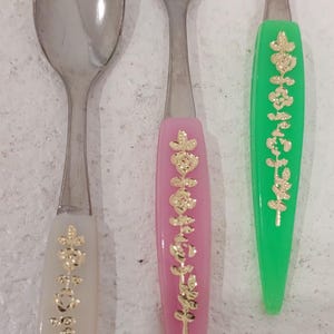 Könnte beinhalten: Drei Löffel mit Edelstahl-Schalen und Griffen in Weiß, Rosa und Grün. Die Griffe haben goldfarbenen Text und florale Designs. Die Löffel sind vertikal angeordnet.