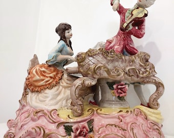 May include: A porcelain figurine of a couple playing music. The woman is seated on a ornate chair and playing a piano. The man is standing and playing a violin. The figurine is decorated with pink, gold, and white details.