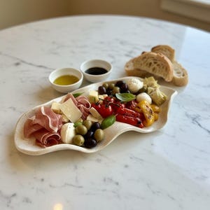 May include: A charcuterie board with various appetizers, including prosciutto, cheese, olives, tomatoes, and roasted peppers. The board is accompanied by small bowls of oil and balsamic vinegar, and slices of bread. The arrangement is on a white, wavy-edged platter.