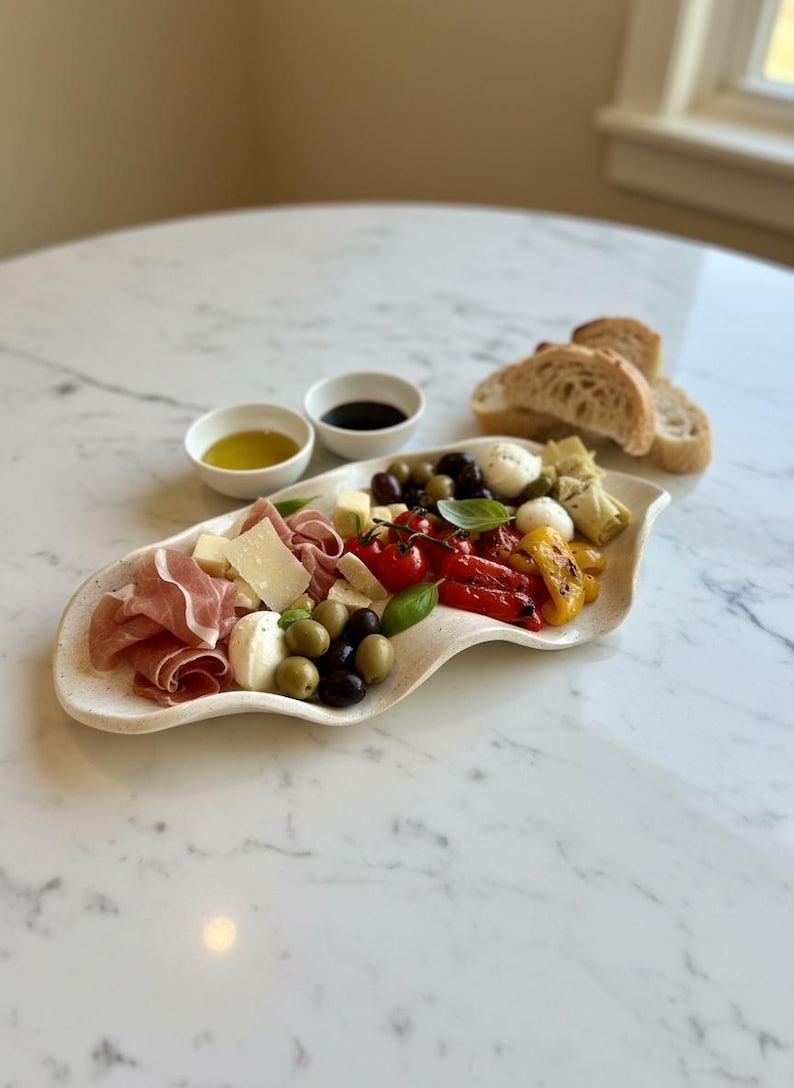 May include: A charcuterie board with various appetizers, including prosciutto, cheese, olives, tomatoes, and roasted peppers. The board is accompanied by small bowls of oil and balsamic vinegar, and slices of bread. The arrangement is on a white, wavy-edged platter.