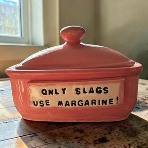 May include: A pink ceramic butter dish with a matching lid and a rectangular inset panel on the front. The panel features the black text "ONLY SLAGS USE MARGARINE!" in a bold, sans-serif font. The dish has a smooth, glossy finish throughout.