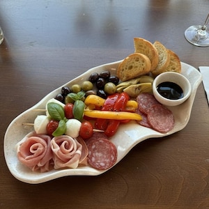 May include: A white, organically shaped platter filled with an assortment of appetizers. The platter includes bread, olives, mozzarella and tomato skewers, salami, prosciutto roses, roasted peppers, artichoke hearts, and a small bowl of balsamic glaze.