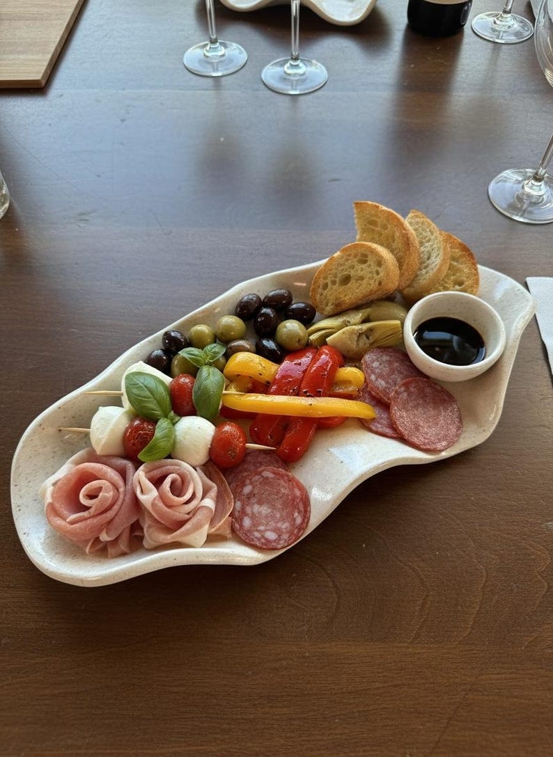May include: A white, organically shaped platter filled with an assortment of appetizers. The platter includes bread, olives, mozzarella and tomato skewers, salami, prosciutto roses, roasted peppers, artichoke hearts, and a small bowl of balsamic glaze.