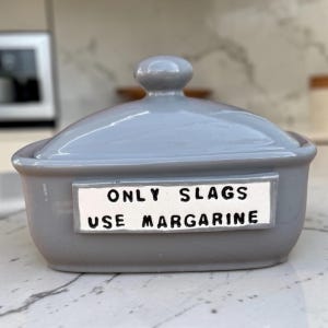 May include: A glossy light grey ceramic butter dish with a matching lid featuring a central knob. A rectangular white plaque is attached to the front, displaying the black text: ONLY SLAGS USE MARGARINE, written in a simple, bold, sans-serif font.