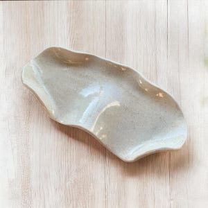 May include: A speckled, off-white ceramic serving dish with a wavy, organic shape. The dish has a glossy finish and is set against a light wood grain background. The design is simple and elegant.