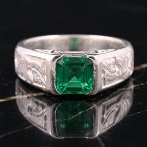 Può includere: Un anello d'argento con una gemma verde smeraldo. La fascia presenta un motivo floreale inciso. L'anello è esposto su una superficie nera riflettente, evidenziando il suo design e il suo colore.
