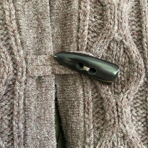 May include: Close-up of a brown cable-knit sweater with a dark brown toggle closure. The sweater has a textured pattern and a button closure. The color is a muted brown.