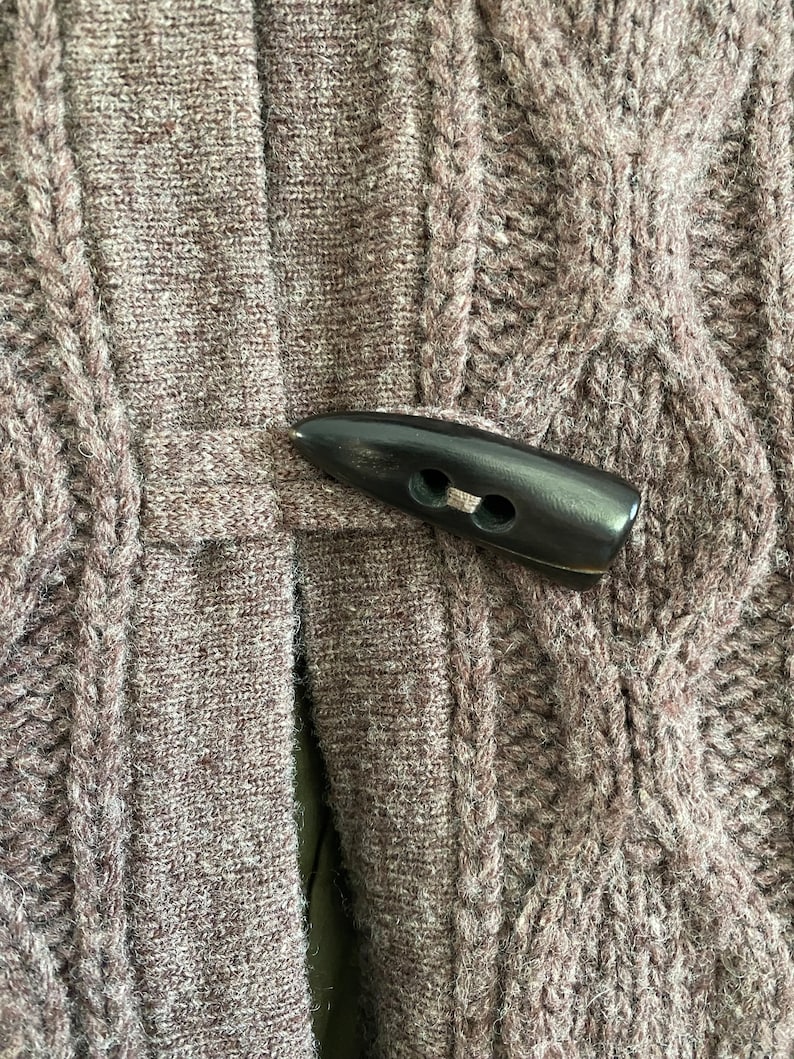 May include: Close-up of a brown cable-knit sweater with a dark brown toggle closure. The sweater has a textured pattern and a button closure. The color is a muted brown.