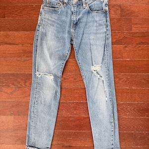 May include: Light blue denim jeans with distressed details, including rips at the knees. The jeans have a classic five-pocket design and a button and zipper closure. The jeans are laid flat on a wooden surface.