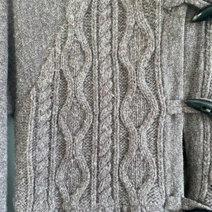 May include: Close-up of a gray knit cardigan with a cable-knit design on the front. The cardigan features a textured pattern and toggle closures. The bottom of the cardigan has a gathered, ruffled hem.