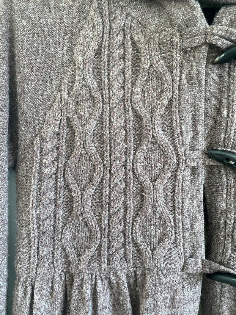 May include: Close-up of a gray knit cardigan with a cable-knit design on the front. The cardigan features a textured pattern and toggle closures. The bottom of the cardigan has a gathered, ruffled hem.