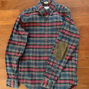 May include: A long-sleeved plaid shirt in a classic pattern of red, green, and navy blue. The shirt features a button-down front, a chest pocket, and brown suede elbow patches. The shirt is made of a soft, flannel-like material.