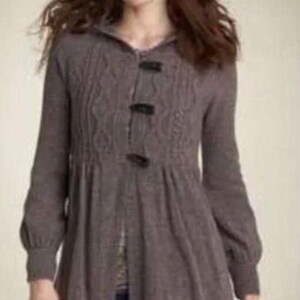 May include: A long, brown knit cardigan with a hooded neckline and three dark buttons. The cardigan features a cable knit design on the upper chest and a gathered waist. The sleeves are long with a slight puff at the shoulders.