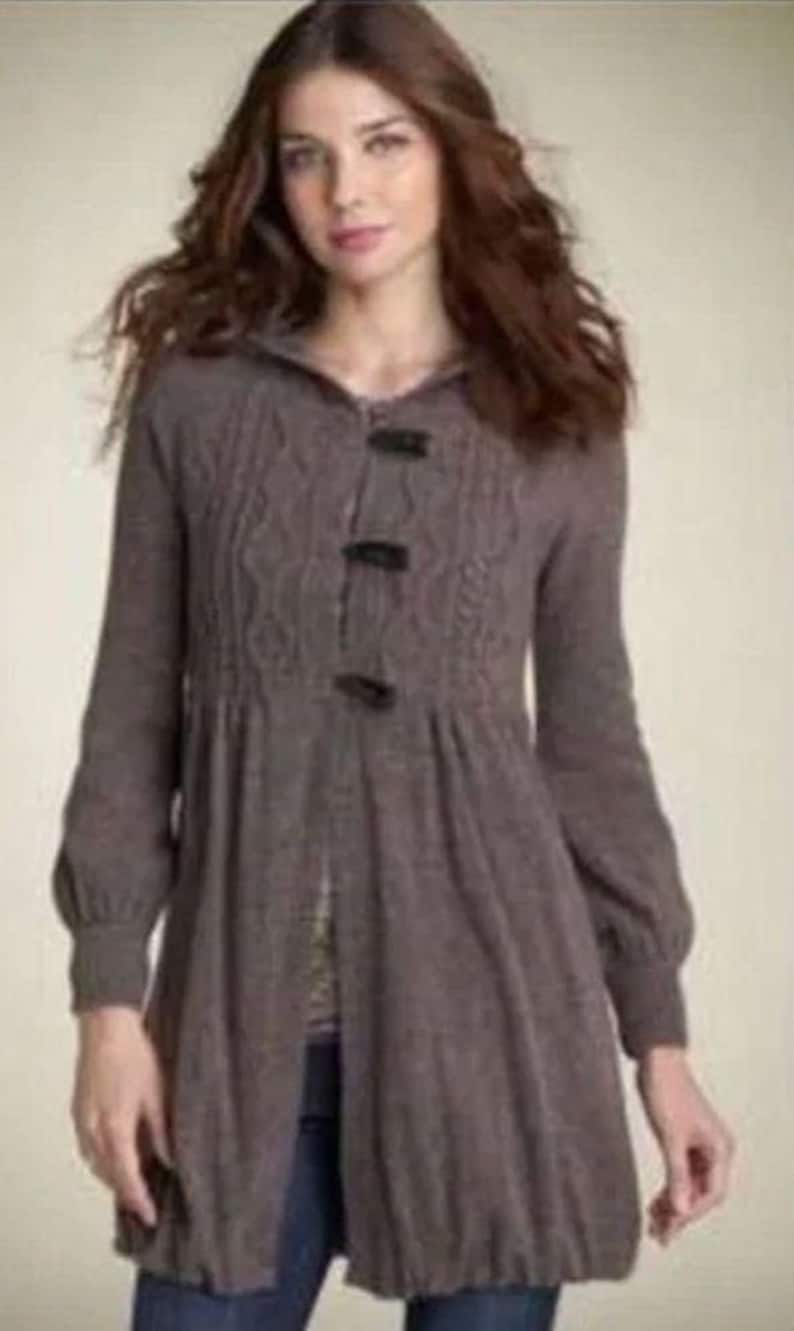 May include: A long, brown knit cardigan with a hooded neckline and three dark buttons. The cardigan features a cable knit design on the upper chest and a gathered waist. The sleeves are long with a slight puff at the shoulders.