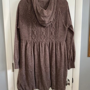 May include: A brown hooded sweater dress with a cable knit design on the back. The dress has long sleeves with ribbed cuffs and a gathered waist. The bottom of the dress has a bubble hem. The dress is hanging on a white door.