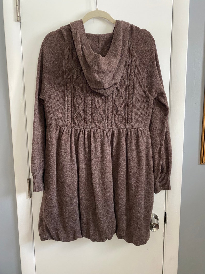 May include: A brown hooded sweater dress with a cable knit design on the back. The dress has long sleeves with ribbed cuffs and a gathered waist. The bottom of the dress has a bubble hem. The dress is hanging on a white door.