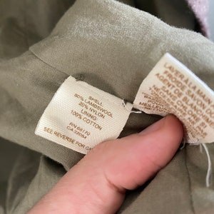 May include: Close-up of a garment's interior, revealing fabric composition and care instructions. The tag indicates an 80% lambswool shell, 20% nylon, and 100% cotton lining. Additional text provides washing instructions. The garment is olive green.