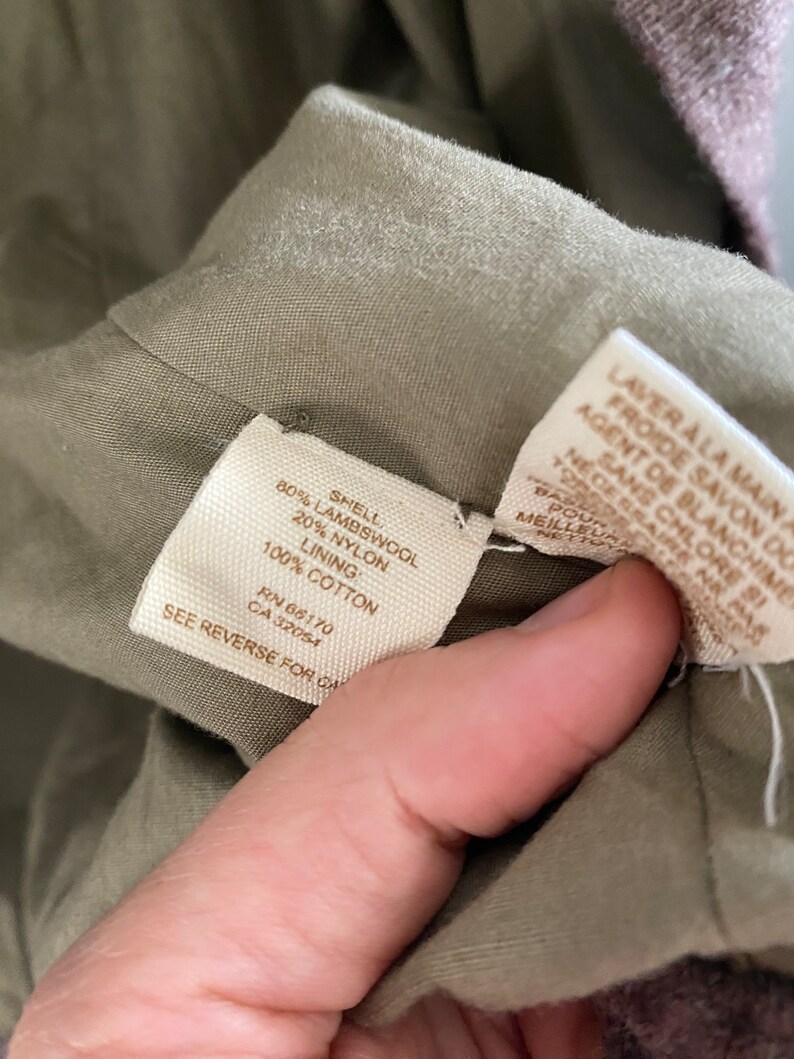 May include: Close-up of a garment's interior, revealing fabric composition and care instructions. The tag indicates an 80% lambswool shell, 20% nylon, and 100% cotton lining. Additional text provides washing instructions. The garment is olive green.