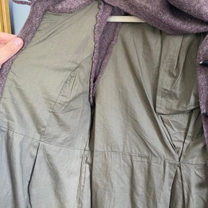 May include: A close-up of a purple-brown coat with a light green lining. The coat is open, revealing the interior lining and stitching details. The coat is hanging on a white hanger.