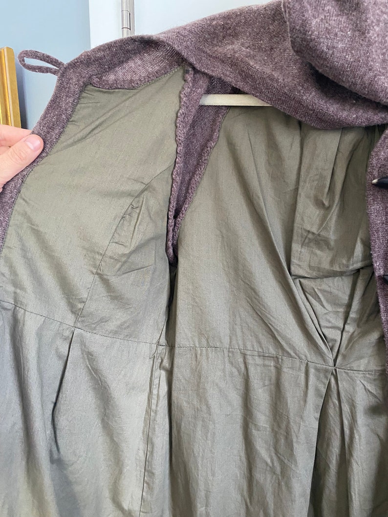 May include: A close-up of a purple-brown coat with a light green lining. The coat is open, revealing the interior lining and stitching details. The coat is hanging on a white hanger.