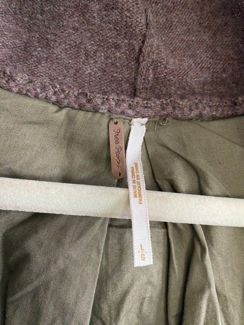 May include: Close-up of a jacket with a brown hood and olive green body. The jacket has a tag that says "Free People" and "MADE IN CHINA FABRIQUE EN CHINE". The jacket is hanging on a white hanger.