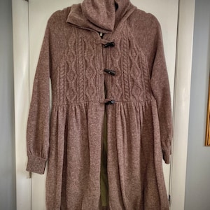 May include: A mauve-colored knit coat with a high collar and long sleeves. The coat features a cable-knit design on the front and is fastened with three dark toggle closures. The bottom of the coat flares out, creating a flowing silhouette.