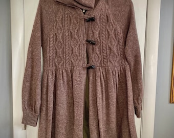 May include: A mauve-colored knit coat with a high collar and long sleeves. The coat features a cable-knit design on the front and is fastened with three dark toggle closures. The bottom of the coat flares out, creating a flowing silhouette.