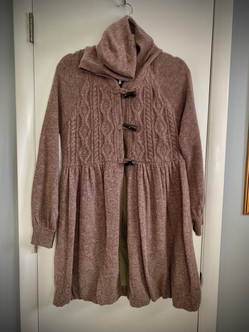 May include: A mauve-colored knit coat with a high collar and long sleeves. The coat features a cable-knit design on the front and is fastened with three dark toggle closures. The bottom of the coat flares out, creating a flowing silhouette.